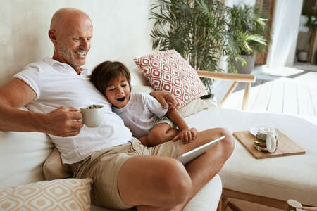 Dad And Son Using Tablet On Family Weekend At Villa. Happy Father With Little Boy Sitting On Sofa, Using Portable Digital Device And Having Fun At Home. Summer Vacation At Tropical Resort.の写真素材