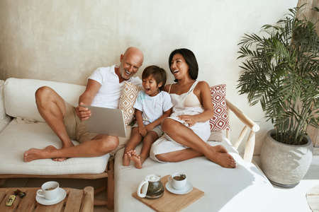 Happy Family On Sofa. Portrait Of Mixed Race Parents With Little Son Using Tablet And Enjoying Leisure On Summer Vacation At Tropical Resort. Different Ethnic Mom And Dad With Boy On Weekend.の写真素材