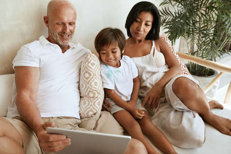 Family With Tablet Portrait. Interracial Parents Using Portable Digital Device Together. Caucasian Father And Asian Mother With Mixed Race Son Sitting On Sofa At Tropical Resort.の写真素材