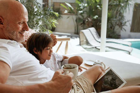 Family Fun At Home. Dad And Little Boy Sitting On Sofa In Tropical Villa Patio With Tablet. Father Drinking Tea And Using Portable Digital Device. Enjoying Leisure On Weekend At Resort.の写真素材