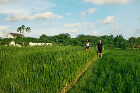 Mature Couple Running On Rice Field In Morning. Caucasian Man And Asian Woman On Jogging Workout Among Green Grass Against Tropical Landscape.の写真素材