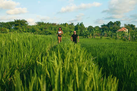 Mature Couple Running On Rice Field In Morning. Caucasian Man And Asian Woman On Jogging Workout Among Green Grass Against Tropical Landscape.の写真素材