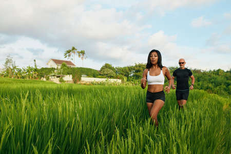 Mature Couple Running On Rice Field In Morning. Caucasian Man And Asian Woman On Jogging Workout Among Green Grass Against Tropical Landscape.の写真素材