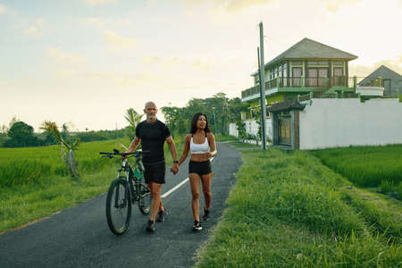 Sportive Couple Walking Bike On Road. Asian Woman And Caucasian Man Walking By Hands With Bicycles And Talking Against Green Tropical Landscape.の写真素材