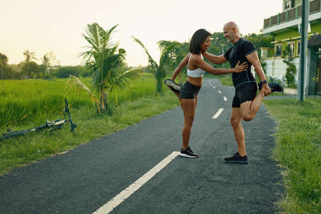 Couple Warming Up Before Running On Road. Asian Woman And Caucasian Man Doing Partner Stretching Before Jogging In Nature.の写真素材