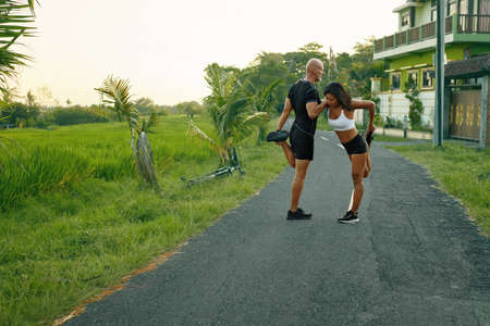 Couple Warming Up Before Running On Road. Asian Woman And Caucasian Man Doing Partner Stretching Before Jogging In Nature.の写真素材