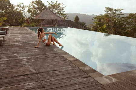Beautiful Girl In Bikini Sitting Near Infinity Pool In Bali, Indonesia. Young Woman With Sexy Body In Swimsuit Portrait.の写真素材