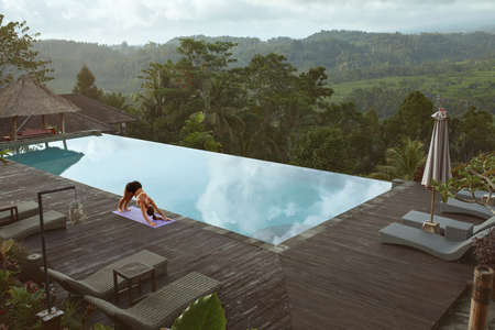 Beautiful Girl Doing Yoga On Poolside In Morning In Bali, Indonesia. Young Slim Woman In Sportswear Standing In Downward Facing Dog Pose On Sport Mat Near Infinity Pool Against Tropical Landscape.の写真素材