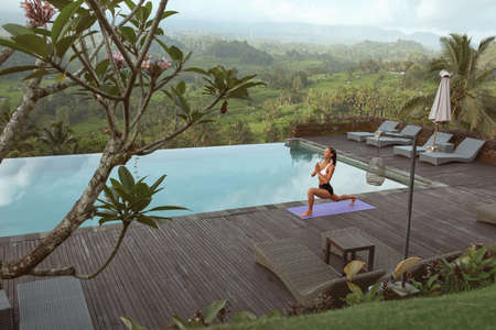 Beautiful Girl Doing Yoga On Poolside In Morning In Bali, Indonesia. Young Woman In Sportswear Standing In Warrior Pose Or Virabhadrasana On Sport Mat Near Infinity Pool Against Tropical Landscape.の写真素材