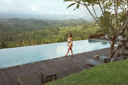 Beautiful Girlâs In Bikini Near Infinity Pool Full-Length Portrait. Young Woman With Sexy Body In White Swimsuit Walks On Wooden Poolside Against Tropical Landscape In Bali, Indonesia.の写真素材