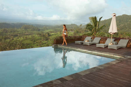 Beautiful Girlâs In Bikini Near Infinity Pool Full-Length Portrait. Young Woman With Sexy Body In White Swimsuit Walks On Wooden Poolside Against Tropical Landscape In Bali, Indonesia.の写真素材