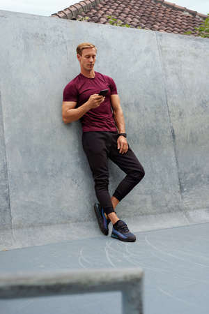 Young Man In Sportswear Using Smartphone Against Concrete Wall. Handsome Caucasian Sportsman With Strong Muscular Body Posing Outdoors.の写真素材
