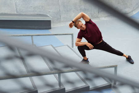 Young Man Stretching Near Concrete Steps Outdoors. Handsome Caucasian Sportsman With Strong Muscular Body In Fashion Sportswear Warming Up Before Intense Workout.の写真素材