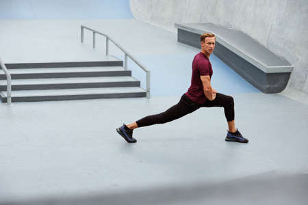 Young Man Stretching Near Concrete Steps Outdoors. Handsome Caucasian Sportsman With Strong Muscular Body In Fashion Sportswear Warming Up Before Intense Workout.の写真素材