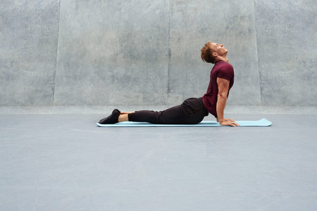 Young Man Stretching On Yoga Mat Against Concrete Wall Outdoors. Handsome Caucasian Sportsman With...