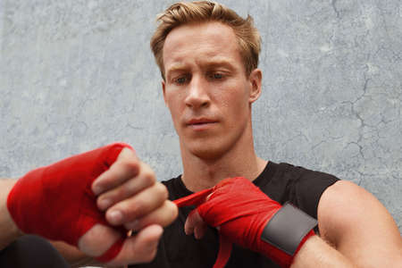 Young Man Wrapping Hand And Wrist With Boxing Tape Against Concrete Wall. Handsome Caucasian Sportsman With Strong Muscular Body In Fashion Sportswear Sitting Outdoors.の写真素材