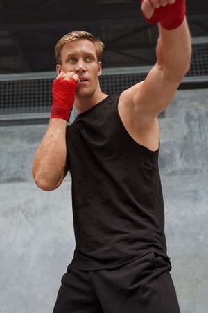 Young Man Stands In Boxing Pose And Doing Punching Workout. Handsome Caucasian Sportsman With Strong Muscular Body In Fashion Sportswear And Hand Wrap On Wrists Against Concrete Wall.の写真素材