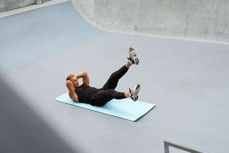 Young Man Doing Abs Workout On Yoga Mat Against Concrete Wall Outdoors. Handsome Caucasian Sportsman With Strong Muscular Body In Fashion Sportswear Warming Up Before Intense Functional Training.の写真素材