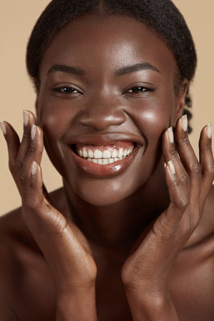 Portrait close up of beautiful african girl. Smiling young woman touch her clean face and looking at camera. Concept of face skin care. Isolated on beige background. Studio shootの写真素材