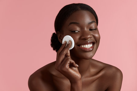 Portrait close up of beautiful black girl touch her clean face with cotton pad. Joyful young woman. Concept of face skin care. Isolated on pink background. Studio shootの写真素材