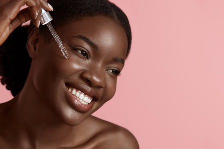 Portrait close up of beautiful black girl dropping serum collagen moisturizer on face. Smiling young woman. Concept of face skin care. Isolated on pink background. Studio shootの写真素材