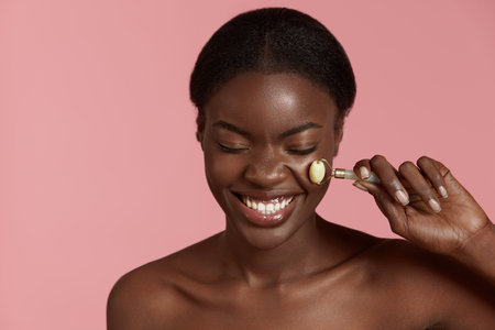 Portrait close up of beautiful african girl massage her face with jade roller. Smiling young woman with closed eyes. Concept of skincare. Isolated on pink background. Studio shootの写真素材