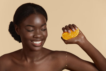 Portrait close up of beautiful african girl pressing slice of orange with drops. Smiling young woman. Concept of skincare. Isolated on beige background. Studio shootの写真素材