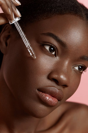 Portrait close up of beautiful black girl dropping serum collagen moisturizer on face. Serious young woman. Concept of face skin care. Isolated on pink background. Studio shootの写真素材