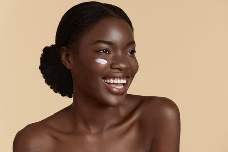 Portrait close up of beautiful black girl with cosmetic cream on face. Smiling young woman with perfect skin. Concept of face skin care. Isolated on beige background. Studio shootの写真素材