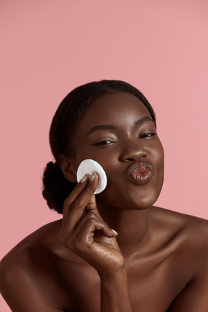 Portrait close up of beautiful black girl touch her clean face with cotton pad. Smiling young woman show her lips. Concept of face skin care. Isolated on pink background. Studio shootの写真素材