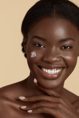 Portrait close up of beautiful black girl with cosmetic cream on face. Smiling young woman looking at camera. Concept of face skin care. Isolated on beige background. Studio shootの写真素材