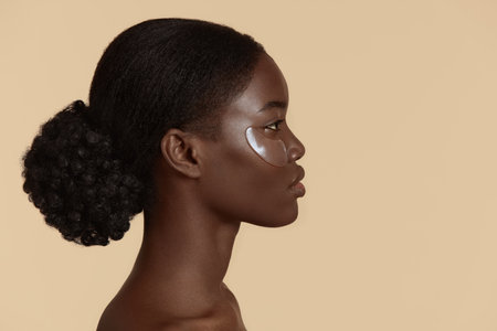 Close up of beautiful african girl with under eye patch on face. Profile of serious young woman with perfect skin. Concept of face skin care. Isolated on beige background. Studio shoot