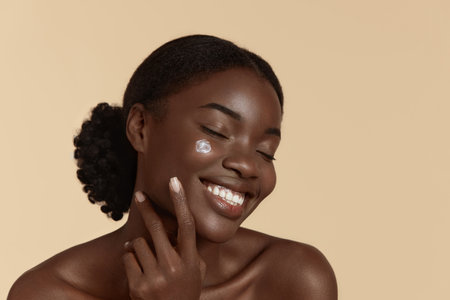 Portrait close up of beautiful black girl with cosmetic cream on face. Smiling young woman with closed eyes. Concept of face skin care. Isolated on beige background. Studio shootの写真素材