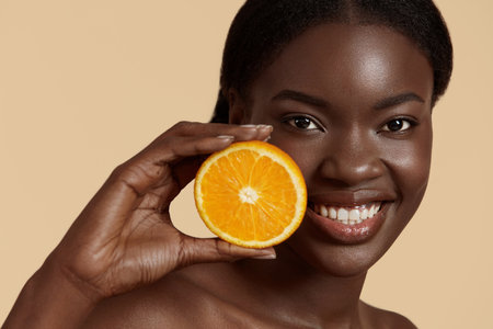 Portrait close up of beautiful african girl hold slice of orange. Smiling young woman looking at camera. Concept of skincare. Isolated on beige background. Studio shootの写真素材