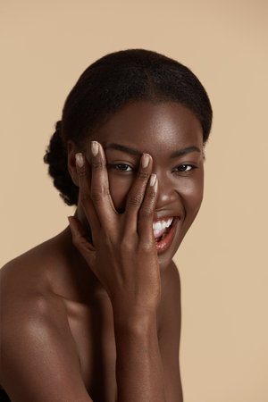 Portrait close up of beautiful african girl. Smiling young woman touch her clean face and looking at camera. Concept of face skin care. Isolated on beige background. Studio shootの写真素材