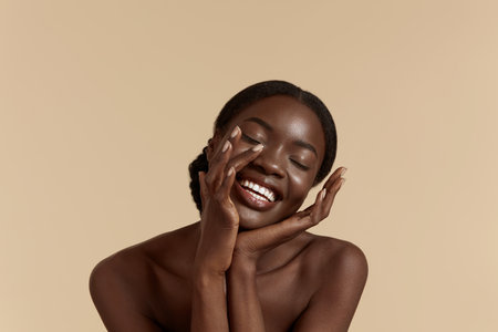Portrait close up of beautiful african girl with closed eyes. Smiling young woman touch her clean face. Concept of face skin care. Isolated on beige background. Studio shootの写真素材