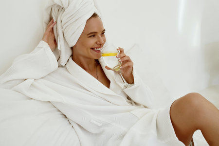 Smiling girl in bathrobe drink lemonade from glass while lay on bed. Young beautiful woman with...