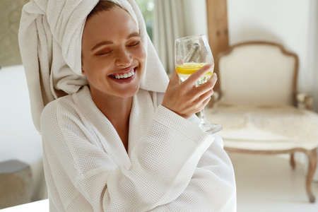 Smiling european girl drink lemonade in bathrobe. Young beautiful woman with wrapped bath towel on head. Concept of rest with spa treatment. Home interiorの写真素材
