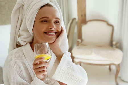 Pleased european girl drink lemonade in bathrobe. Young smiling beautiful woman with wrapped bath towel on head. Concept of rest with spa treatment. Home interiorの写真素材