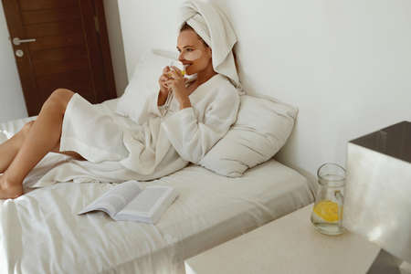 Pleased european girl lay on bed and drink lemonade. Young beautiful woman with under eye patch on face wear bathrobe and wrapped bath towel on head. Concept of rest. Home bedroom interiorの写真素材