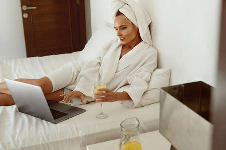 European girl use laptop and drink lemonade while lay on bed. Young pretty woman with under eye patch on face wear bathrobe and wrapped bath towel on head. Concept of rest. Home bedroom interiorの写真素材