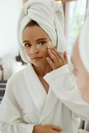 European girl applying under eye patch on face and watch in mirror. Young focused woman eye wear bathrobe and wrapped bath towel on head. Concept of face skin careの写真素材