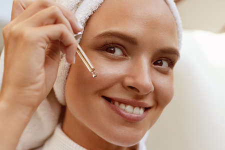 Girl dropping serum collagen moisturizer from pipette on face close up. Young beautiful woman with wrapped bath towel on head. Concept of face skin careの写真素材