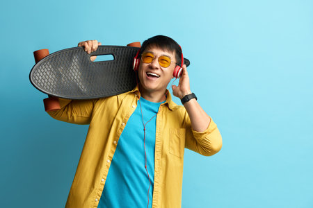 Asian Man Listening Music. Positive Man Wearing Headphones Carrying Longboardの写真素材