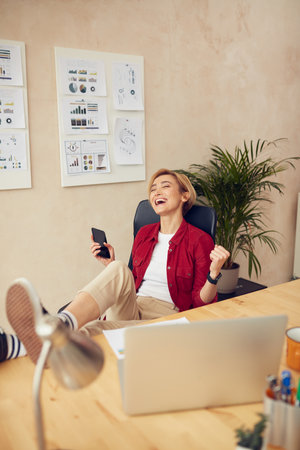 Business Woman Celebrate At Office. Cheerful Woman Holding Mobile And Feeling Happyの写真素材