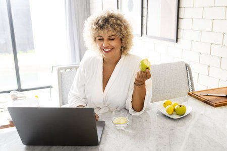 Smiling Woman in Bathrobe Enjoying Healthy Breakfast and Laptopの写真素材