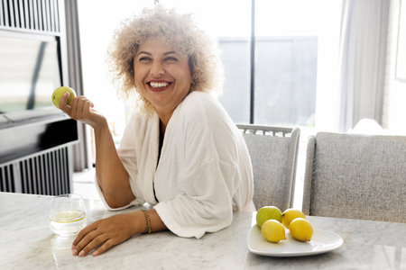 Happy Mature Woman with Apple Enjoying Morning in Bright Kitchenの写真素材