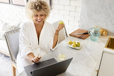 Woman in Bathrobe Working From Home with a Laptop and Snackingの写真素材