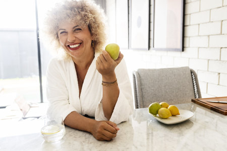 Cheerful Woman in White Robe Holding Apple in Sunny Kitchenの写真素材