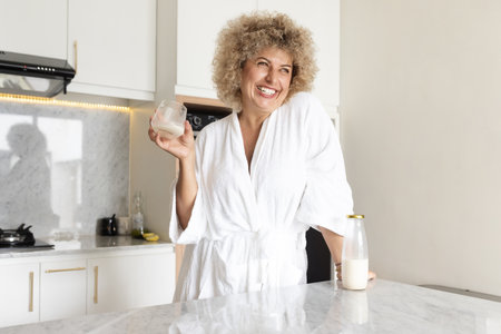 Happy Woman in Robe Enjoying a Glass of Milk in Kitchenの写真素材